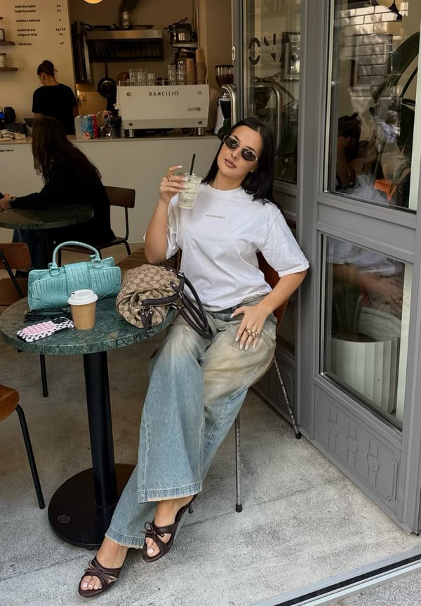 Vrouw met zonnebril drinkt een ijskoud drankje terwijl ze aan een cafétafel zit met twee handtassen, een koffiekop en een telefoon.