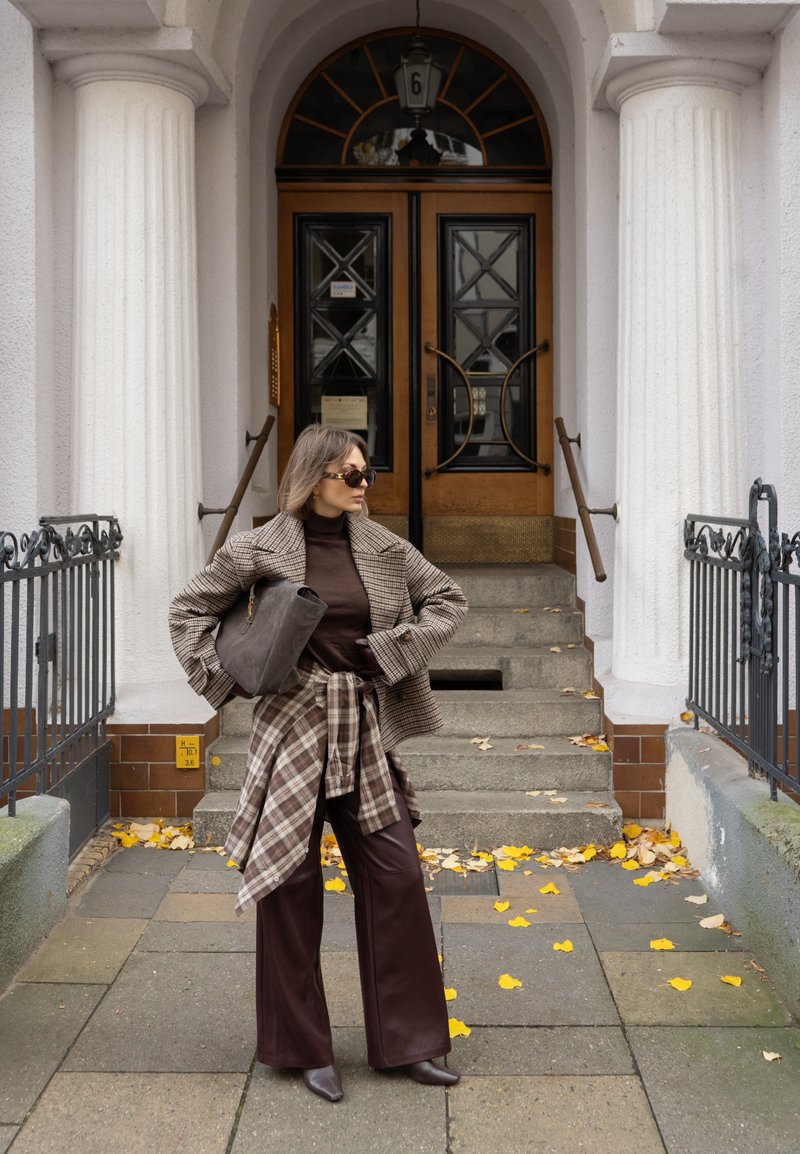 Blazer xadrez castanho sobre uma camisa de gola alta castanha, calças largas de couro castanho e mala combinando. Camisa xadrez decorativa atada à cintura.