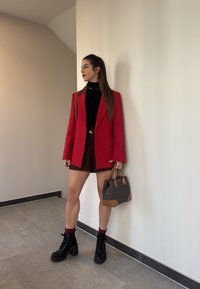 Rød blazer, sort turtleneck, glitrende shorts og sorte snørebåndsstøvler. Brun håndtaske med detaljer. Står imod en hvid væg.