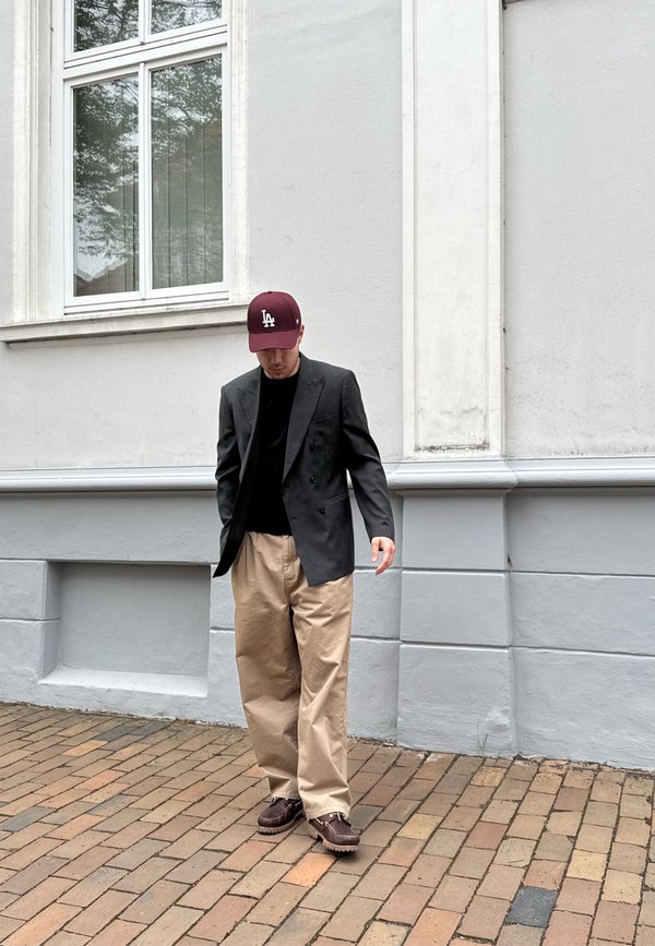 Muž nosiaci bordovú čiapku LA, čierny blazer, čiernu košeľu, béžové široké nohavice a hnedé topánky stojí na tehličkovom chodníku pri šedej budove.