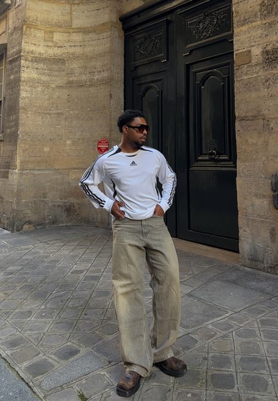 Hombre con camiseta blanca de manga larga de Adidas, pantalones anchos beige y gafas de sol, de pie con las manos en las caderas en una calle adoquinada cerca de una puerta negra.