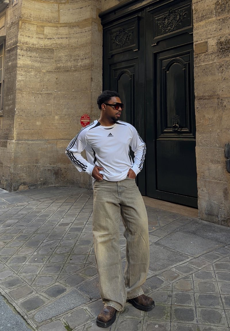 Mees, kes kannab valget Adidas pika varrukaga särki, beežisid laia säärega pükse ja päikeseprille, seisab käed puusadel munakiviteel musta ukse lähedal.