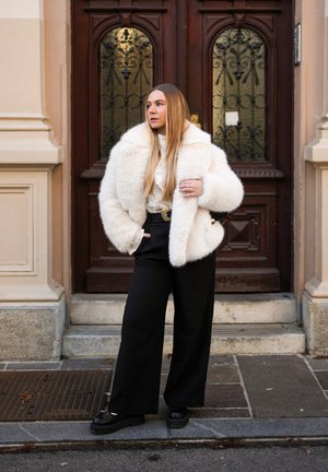 Abrigo de piel sintética blanco, tela texturizada, pantalones negros anchos, zapatos de plataforma gruesos, de pie frente a una puerta de madera ornamentada con cristal.