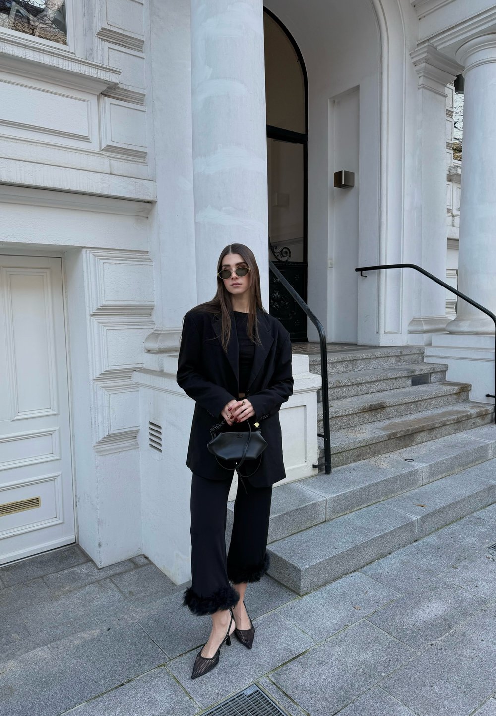 Μαύρο φαρδύ σακάκι πάνω από μαύρο τοπ, συνδυασμένο με μαύρο παντελόνι μέχρι τον αστράγαλο με γούνινο τελείωμα. Αξεσουάρ: στρογγυλά γυαλιά ηλίου και μικρή τσάντα χειρός.