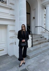 Μαύρο φαρδύ σακάκι πάνω από μαύρο τοπ, συνδυασμένο με μαύρο παντελόνι μέχρι τον αστράγαλο με γούνινο τελείωμα. Αξεσουάρ: στρογγυλά γυαλιά ηλίου και μικρή τσάντα χειρός.