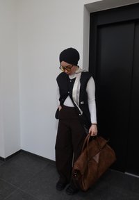 Femme portant un turban noir, des lunettes de soleil, un gilet noir, une chemise blanche à manches longues et un pantalon marron, tenant un grand sac en cuir marron près de la porte de l'ascenseur.