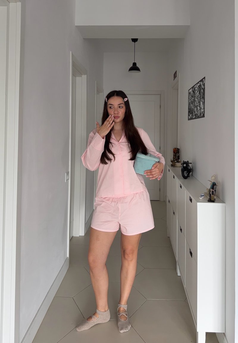 Femme en pyjama rose se tient dans le couloir, tenant une petite boîte bleue et se couvrant la bouche avec une main.