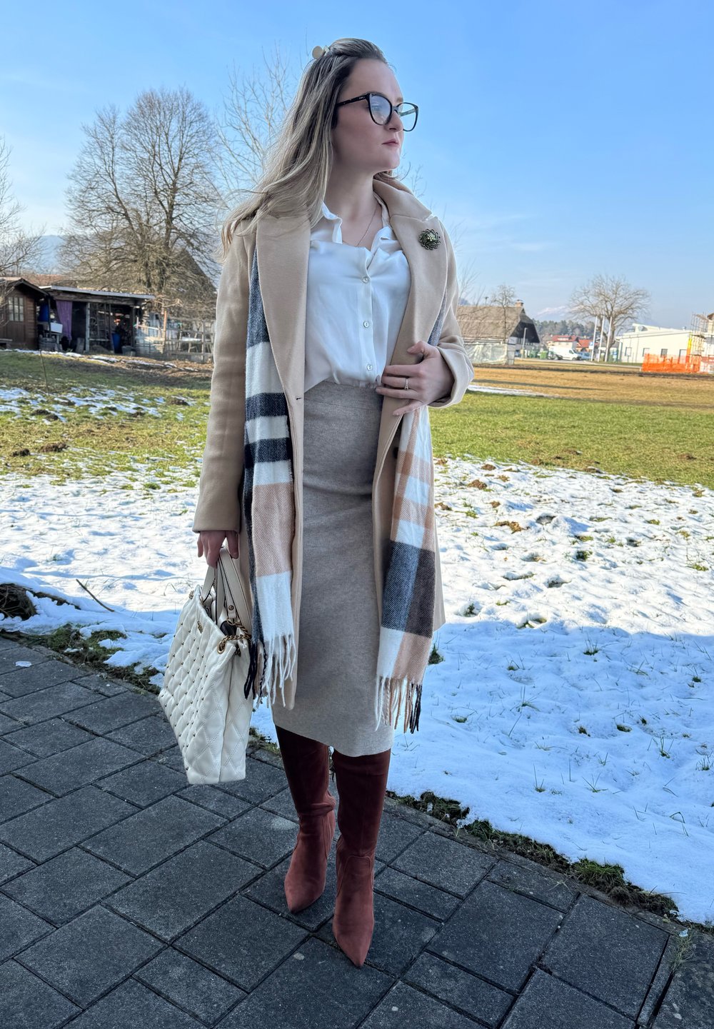 Casaco bege, blusa branca, saia bege, cachecol estampado com franjas, bolsa branca e botas burgundy até ao joelho numa calçada coberta de neve.