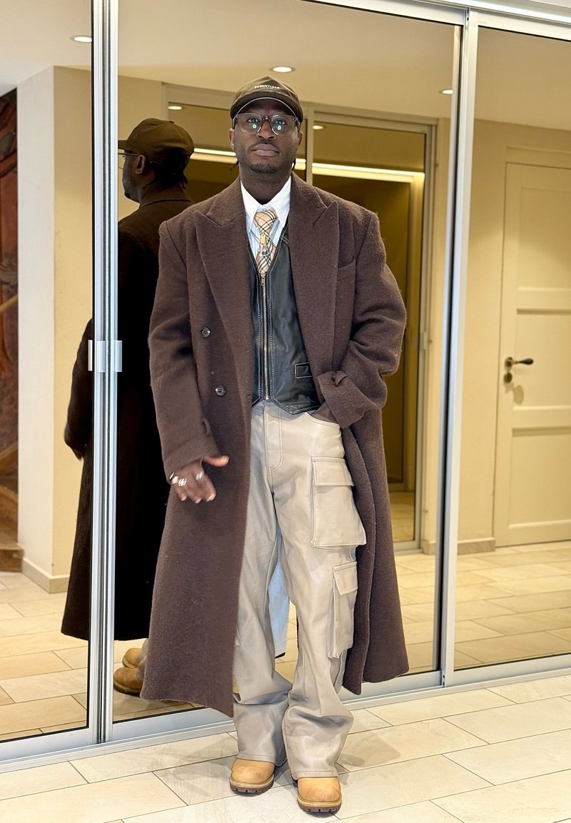 Homme portant un long manteau marron, un gilet en cuir noir, une cravate à carreaux, un pantalon cargo beige, des bottes couleur sable, des lunettes et une casquette noire, debout à l'intérieur près de portes vitrées.