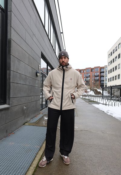 Hombre con chaqueta beige con cremallera, gorro gris, pantalones negros y zapatillas, de pie sobre un pavimento mojado con nieve y edificios modernos al fondo.
