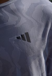 Helles graues Sportshirt mit strukturiertem Muster, Perforationen und einem schwarzen Logo-Akzent auf der oberen linken Seite.
