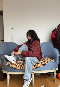 Γυναίκα με σγουρά μαλλιά φοράει μπορντό μπουφάν και ανοιχτόχρωμο τζιν δένει λευκά αθλητικά παπούτσια ενώ κάθεται σε μπλε ρετρό καναπέ με μοτίβο κουβέρτα.