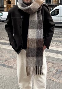Man wearing a dark coat, a large fuzzy checkered scarf in brown and grey tones, and light-coloured trousers, standing on a city street.