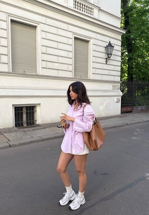 Camisa de rayas rosa claro de corte oversized, pantalones cortos blancos de encaje, zapatillas gris y un bolso tote de cuero marrón. Acera con una superficie texturizada gris.