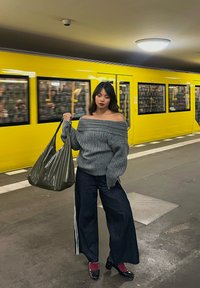 Oversized grey knitted jumper, wide-leg dark denim trousers with side stripes, glossy green handbag, black patent heels, set in a subway station.