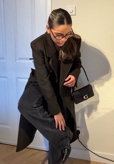 Abrigo largo negro, pantalones negros ajustados con textura brillante y un pequeño bolso negro con un detalle dorado. Ambiente interior con una puerta visible.