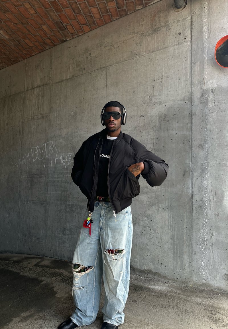 Homme portant une doudoune noire, un jean oversized avec de grandes déchirures, des lunettes de soleil noires, un casque audio, debout contre un mur en béton gris avec des graffitis.