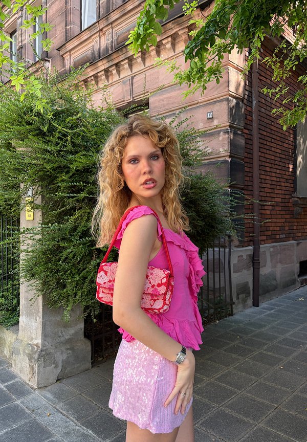 Giovane donna con capelli biondi ricci, con top rosa con volant e gonna scintillante, in piedi all'aperto vicino a vegetazione e vecchio edificio in mattoni.