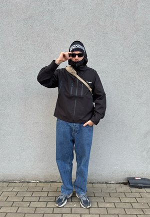 Chaqueta negra impermeable con bolsillos con cremallera, jeans azules de corte holgado, zapatillas grises y un gorro de punto negro con letras blancas.