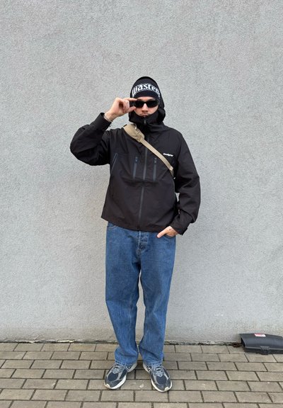 Chaqueta negra impermeable con bolsillos con cremallera, jeans azules de corte holgado, zapatillas grises y un gorro de punto negro con letras blancas.