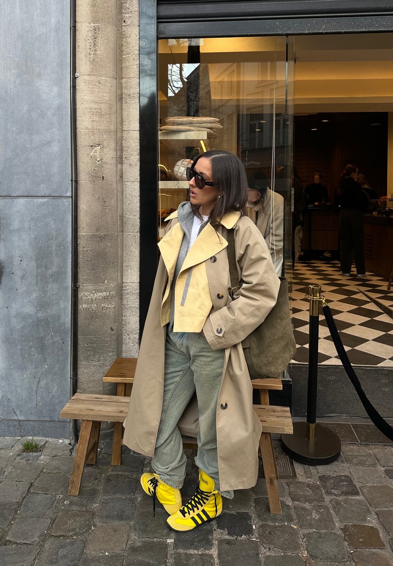 Trench-coat beige sur une veste courte jaune clair, associé à un pantalon gris et des baskets Adidas jaune vif. Sac en daim texturé. Lunettes de soleil noires.