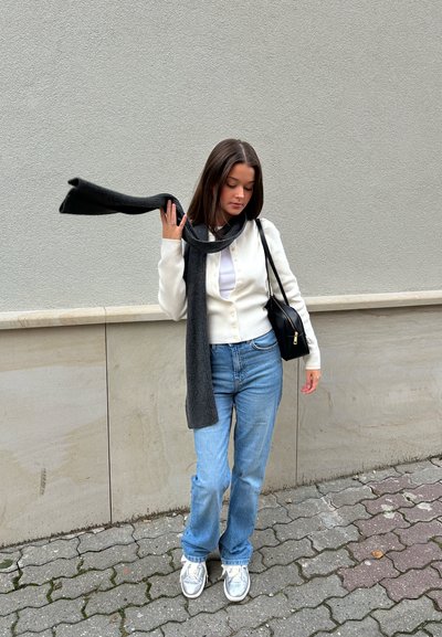 Cárdigan blanco de botones sobre una blusa blanca, combinado con jeans azul claro y zapatillas plateadas. Una bufanda gris oscuro cuelga alrededor del cuello. Bolsa negra.