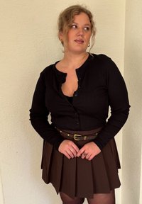 Camisa negra de manga larga acanalada con un escote abotonado, superpuesta sobre una camiseta ajustada, combinada con una falda marrón con pliegues y un cinturón marrón.