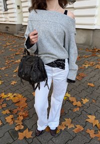 Pull gris surdimensionné, superposé avec une chemise en dentelle noire, pantalon blanc à jambes larges, sac à main sombre avec des pampilles, et chaussures marron, debout sur un chemin jonché de feuilles.