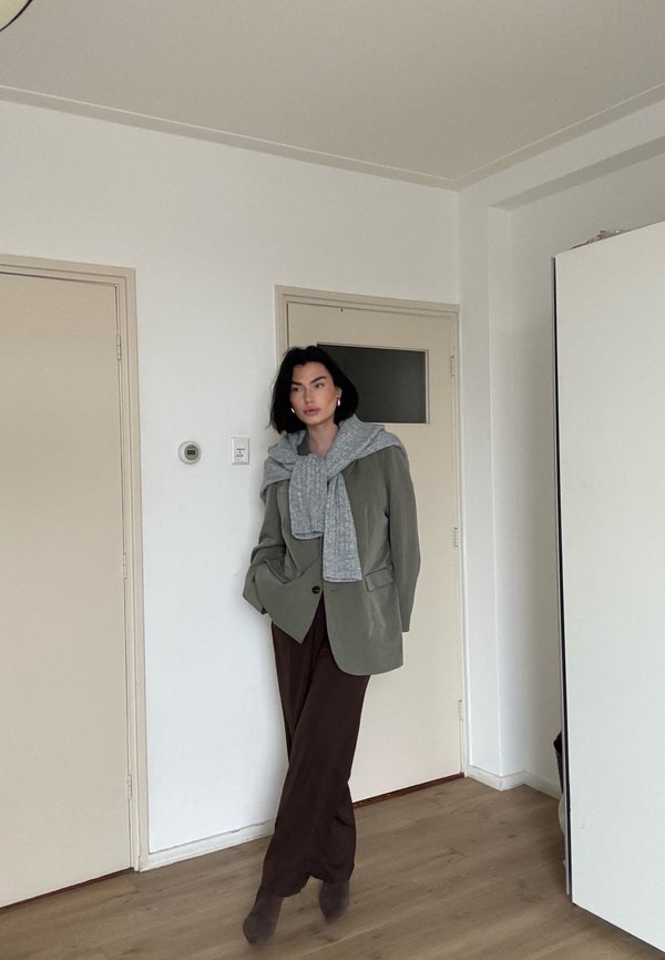 Kvinde der står indendørs iført en olivengrøn blazer, grå sweater lagt over skuldrene, brune bukser og ankelstøvler, lænende sig mod en lukket dør.