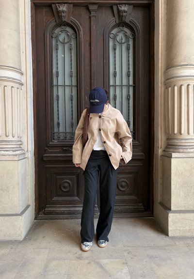 Persona de pie frente a una puerta de madera ornamentada, vistiendo una chaqueta beige, pantalones negros de rayas finas, zapatos plateados y gorra azul marino, mirando hacia abajo.