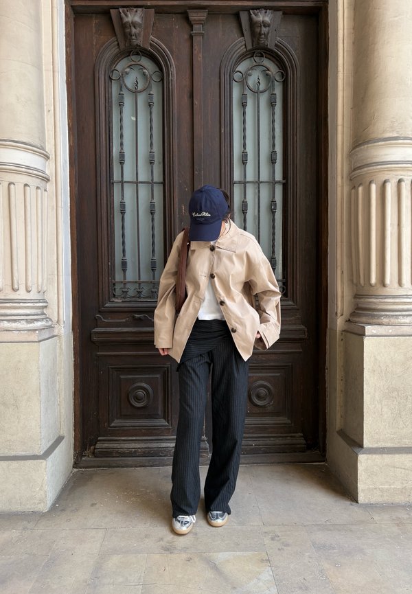 Osoba stojąca przed ozdobionymi drewnianymi drzwiami, ubrana w beżową kurtkę, czarne spodnie w cienkie paski, srebrne buty i granatową czapkę, patrząca w dół.