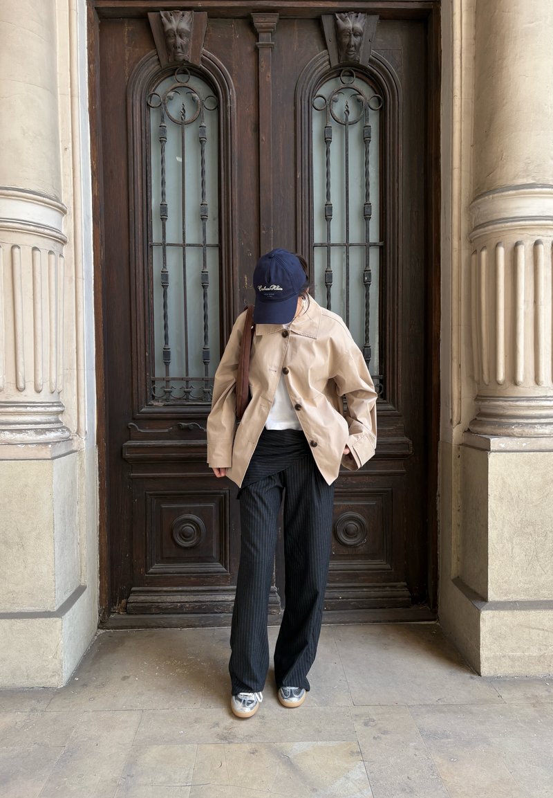 Personne se tenant devant une porte en bois ornée, portant une veste beige, un pantalon noir à fines rayures, des chaussures argentées et une casquette bleu marine, regardant vers le bas.