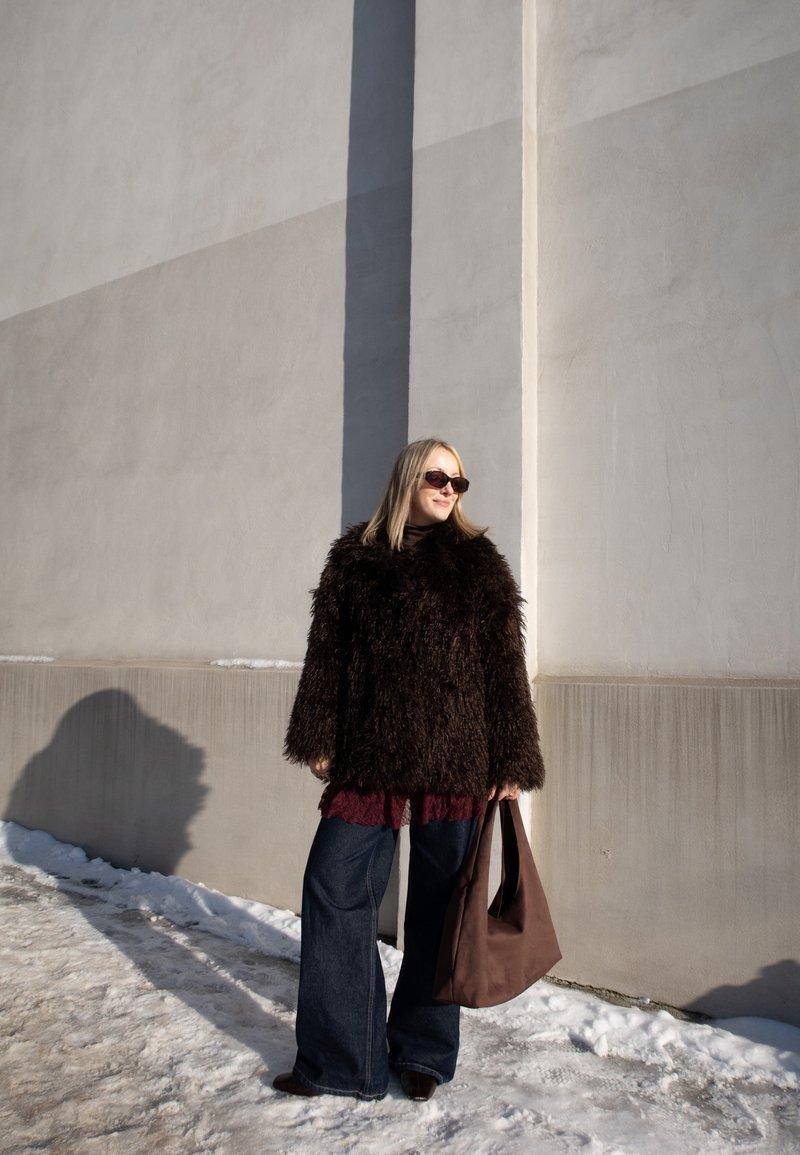 Femme portant un manteau en fourrure foncé et des lunettes de soleil, debout sur un sol enneigé près d'un mur beige ensoleillé, tenant un grand sac marron.