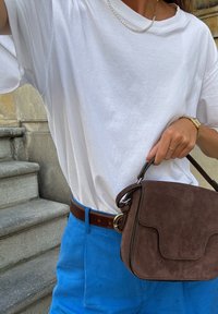 Bolsa de camurça castanha com fecho de aba, combinada com uma t-shirt de algodão branca, calções de linho azuis e um cinto texturizado castanho.