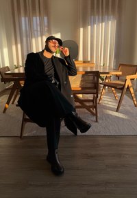 Μαύρο διπλό αλπικό παλτό, μαύρο πουκάμισο και μαύρες μπότες. Ξύλινο τραπέζι φαγητού και καρέκλες στο βάθος, απαλός φυσικός φωτισμός.