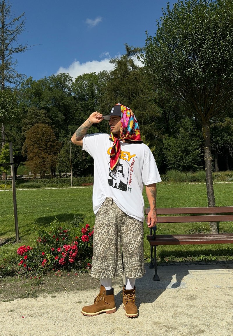 Λευκό γραφικό T-shirt με λογότυπο "Obey", κοντά παντε shorts με λεοπάρ σχέδιο, καφέ μπότες, πολύχρωφο κασκόλ, σε πάρκο.