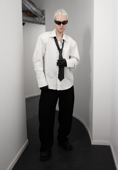 Persona con cabello rubio corto, llevando gafas de sol negras, camisa blanca de gran tamaño, corbata y guantes negros, pantalones negros, de pie en un pasillo estrecho y curvo.