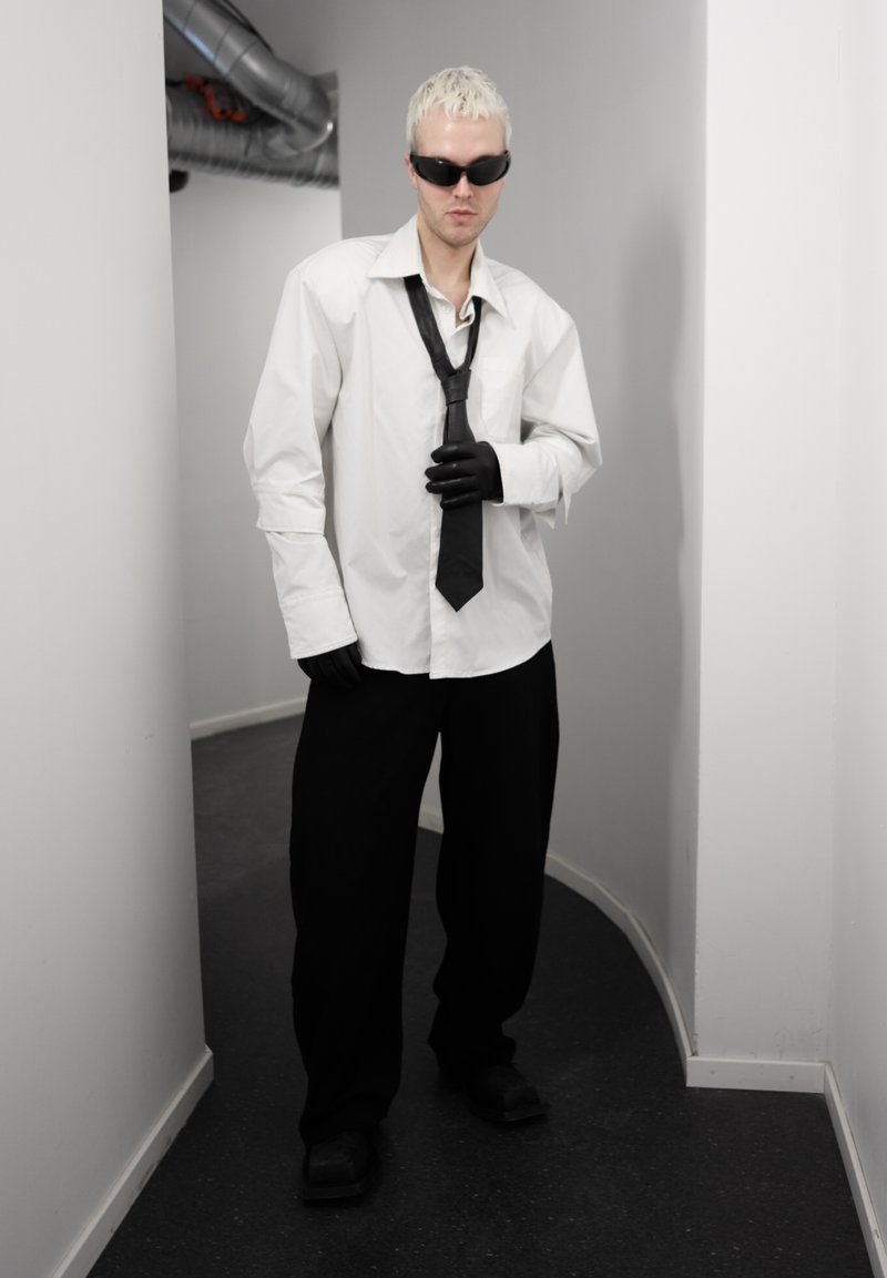 Personne aux cheveux blonds courts portant des lunettes de soleil noires, une chemise blanche oversize, une cravate noire et des gants, un pantalon noir, debout dans un couloir étroit et courbé.