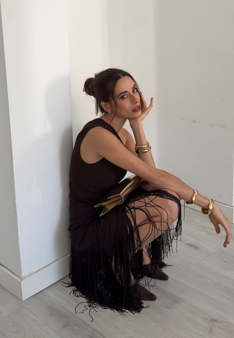 Femme en robe noire à franges assise sur ses talons contre un mur blanc, tenant une pochette dorée, portant des bracelets dorés et des bottines.