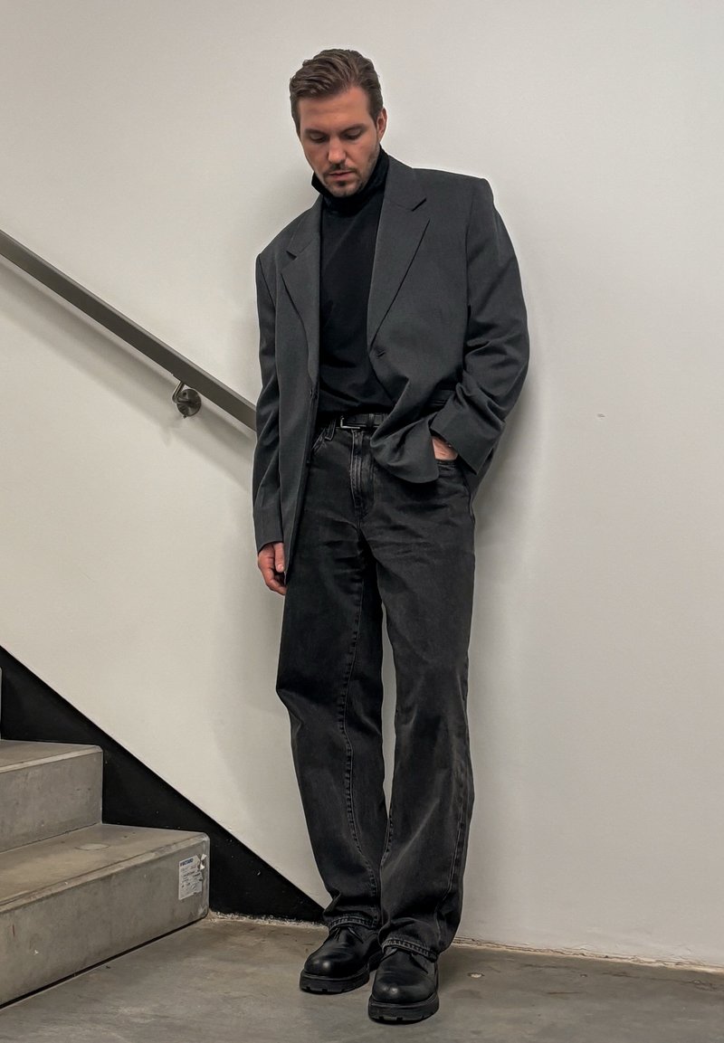 Homme appuyé contre un mur blanc près des escaliers, portant un col roulé noir, un blazer gris oversize, un jean noir et des chaussures noires.