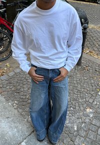 Langärmliges weißes Baumwoll-T-Shirt kombiniert mit weiten, hellblauen Jeans aus Denim. Die Jeans haben eine ausgewaschene Textur und sichtbare Nähte.
