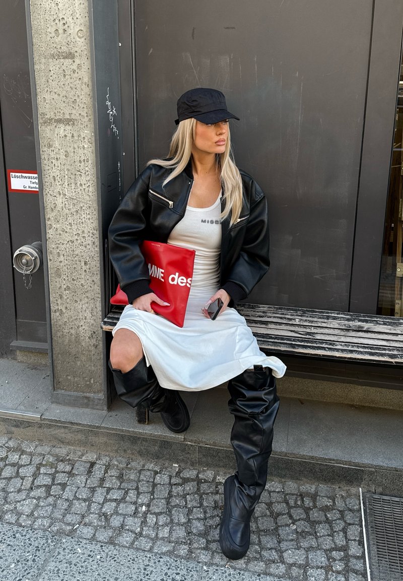 Noor naine musta bareti, nahktagi, valge kleidi ja põlveni ulatuvate mustade saapastega istub pingil, hoides punast käekotti ja telefoni.