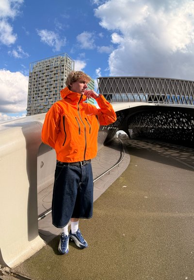 Chaqueta impermeable naranja con bolsillos con cremallera, combinada con pantalones cortos de mezclilla azul oscuro. Las zapatillas son azules con acentos blancos, usadas con calcetines blancos.
