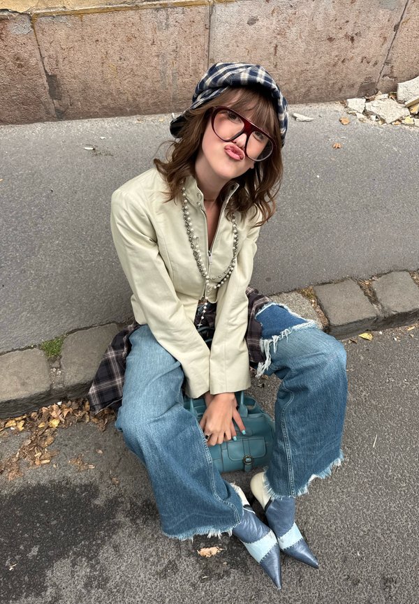Joven sentada en la acera con sombrero de cuadros, gafas grandes, chaqueta ligera, vaqueros anchos rotos y zapatos puntiagudos bicolor, sujetando una bolsa.