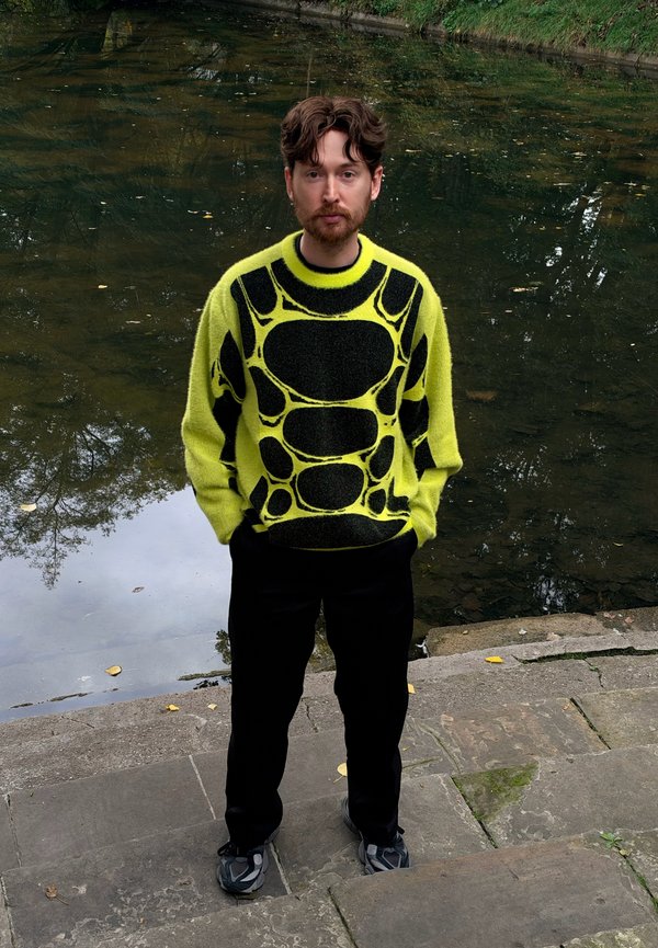 Hombre de pie en escalones de piedra junto al agua, vistiendo un suéter con estampado en amarillo neón y negro, con pantalones negros y zapatillas deportivas.