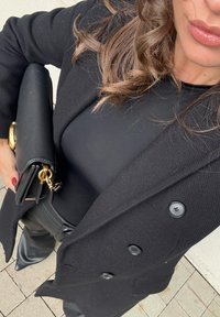 Schwarzer, strukturierter Mantel mit doppelten Knöpfen, kombiniert mit einem taillierten schwarzen Oberteil und glänzenden schwarzen Hosen. Eine schwarze Clutch mit goldener Kettendetail.