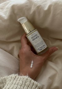 Klares Glasfläschchen mit goldfarbenem Pumpspender, beschriftet mit "Good Genes Glycolic Acid Treatment." Weiße Creme auf der Hand, weicher beige Stoffhintergrund.