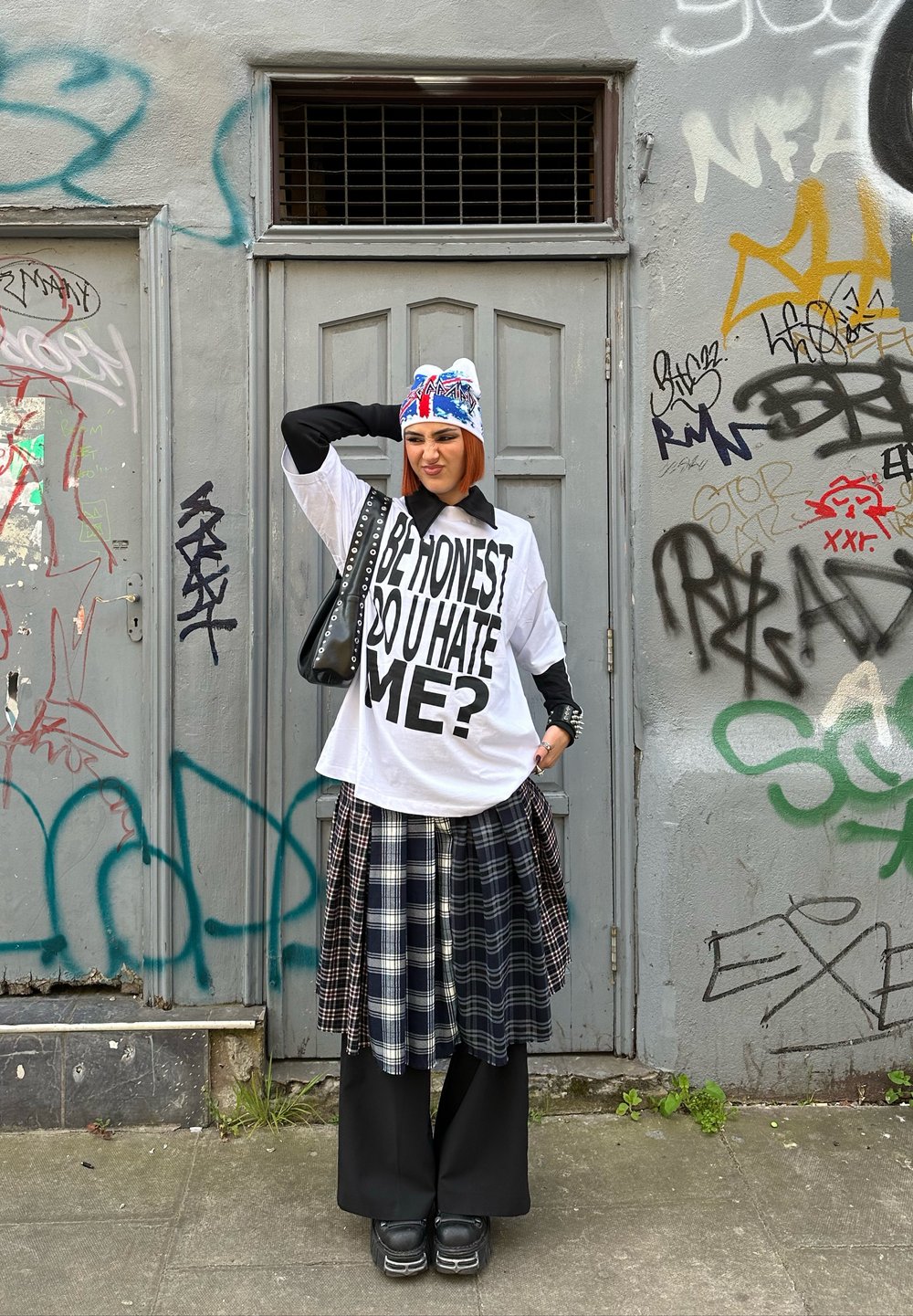 T-shirt bianco oversize con testo nero, sovrapposto a una gonna a quadri in tonalità blu navy e grigie, abbinato a pantaloni larghi neri e scarpe nere chunky.