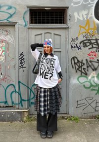 T-shirt bianco oversize con testo nero, sovrapposto a una gonna a quadri in tonalità blu navy e grigie, abbinato a pantaloni larghi neri e scarpe nere chunky.