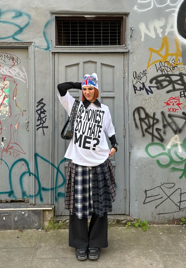 T-shirt bianco oversize con testo nero, sovrapposto a una gonna a quadri in tonalità blu navy e grigie, abbinato a pantaloni larghi neri e scarpe nere chunky.